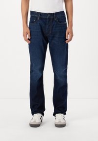 Jeans din denim bleumarin închis, cu croi drept, având buzunare frontale și închidere clasică cu nasture și fermoar. Purtați cu adidași albi.