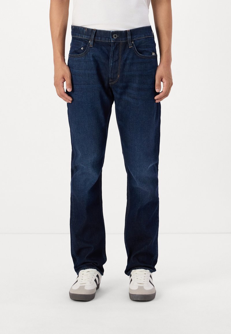 Jeans din denim bleumarin închis, cu croi drept, având buzunare frontale și închidere clasică cu nasture și fermoar. Purtați cu adidași albi.