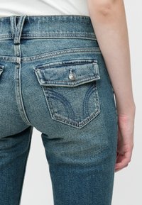 Tuvojums aizmugures kabatai uz ziliem džinsiem ar atloku un sudraba pogu, kuru valkā persona ar baltu augšu uz vienkrāsaina fona.
