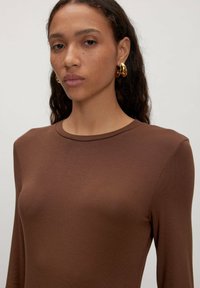 Frau mit lockigem dunklem Haar, die ein braunes Langarmoberteil und große goldene Creolen trägt, neutraler Hintergrund.