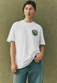 Hvid bomulds t-shirt med et grønt grafisk logo på venstre bryst, parret med blå denim jeans. T-shirten har en afslappet pasform.
