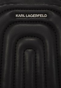 Musta nahkatuote, jossa on tekstuurisia konsentrisia kuvioita, ommeltuja yksityiskohtia sekä hopeinen logo, jossa lukee "KARL LAGERFELD" ylhäällä.