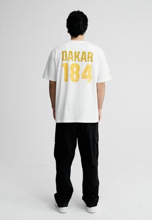 Mężczyzna ubrany w biały t-shirt z dużym żółtym napisem "DAKAR 184" z tyłu, czarne spodnie typu cargo i białe trampki, stoi na jednolitym tle.