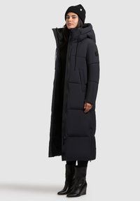 Donna che indossa un lungo cappotto imbottito nero con cappuccio, un berretto nero e stivali con zeppa neri, in piedi di fronte a uno sfondo semplice.