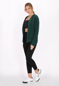 Giacca in pile verde con zip e cappuccio, abbinata a un top corto nero e leggings neri. Indossata con sneakers bianche con accenti neri.