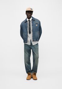 Νεαρός άνδρας που στέκεται, φοράει μπεζ καπέλο, μπλε τζιν μπουφάν, λευκό πουκάμισο με μαύρη γραβάτα, ξεθωριασμένο μπλε τζιν και καφέ μπότες εργασίας.