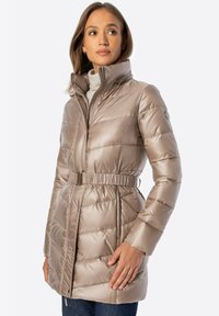 Mujer con abrigo acolchado beige con cinturón, cuello alto y cremallera, combinada con un suéter de cuello alto gris claro y pantalones azul oscuro.