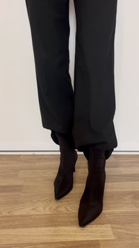 Crne gležnjače s visokim potpeticama, špicastim vrhom i glatkim, teksturiranim završetkom, uparene s širokim crnim hlačama, stoje na drvenom podu.