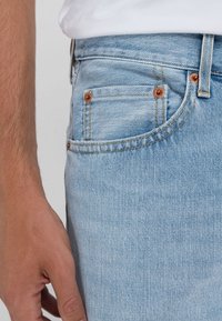 Närbild på en ljusblå jeansficka i denim med synliga kopparnitar och en avslappnad hand bredvid, iförd en vit skjorta.