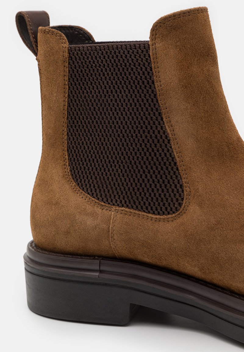 Bottine Chelsea en suede marron avec panneau latéral en élasthanne sombre et semelle noire à faible talon, vue de côté en gros plan.