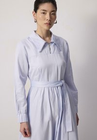 Robe bleu clair avec un grand col, détails zippés, manches longues et taille ceinturée. Tissu lisse et design de jupe froncée.