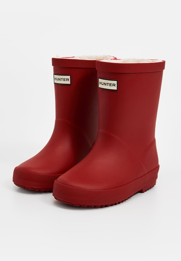 Bottes de pluie en caoutchouc rouge avec une finition lisse, doublure en fausse fourrure et semelle texturée. Présente un logo rectangulaire sur le côté extérieur.