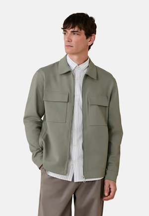 Mand med kort mørkt hår iført en lysegrøn lynlåstrøje over en hvidstribet skjorte og taupefarvede bukser, der kigger til siden.