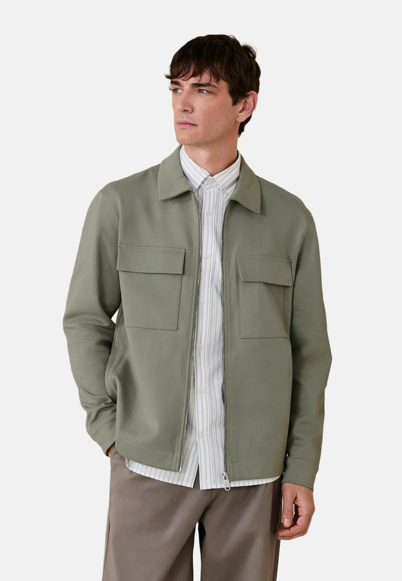 Homme aux cheveux courts et foncés portant une veste zippée vert clair sur une chemise blanche à rayures boutonnée et un pantalon taupe, regardant sur le côté.