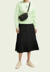 Felpa verde chiaro con scritta, gonna midi a pieghe nera e sneakers beige con dettagli pesca; borsa a tracolla nera con accessori dorati.