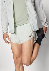 Pantalones cortos deportivos de color verde claro y una camiseta ajustada a juego, cubierta con una chaqueta ligera reflectante. Calcetines oscuros y zapatillas negras con suelas texturizadas.