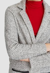 Grauer strukturierter Blazer mit Reverskragen und einem schwarzen Taschen-Detail, getragen über einem roten Rollkragenpullover. Verfügt über eine taillierte Passform.