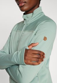 Helle grüne Fleecejacke mit Reißverschluss und strukturiertem Finish, mit einem dekorativen Emblem am Ärmel und Daumenlöchern in den Bündchen.