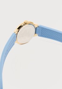 Πίσω πλευρά χρυσού ρολογιού χειρός Tommy Hilfiger με ανοιχτό μπλε λουρί από υφή καουτσούκ και χαραγμένες λεπτομέρειες της μάρκας.