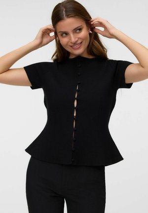 Jeune femme aux longs cheveux bruns portant un haut noir ajusté à manches courtes avec des boutons sur le devant et un pantalon noir, ajustant ses cheveux et souriant.