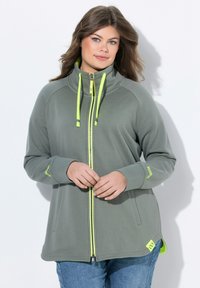 Chaqueta gris con cremallera y cuello alto, adornada con acentos en amarillo neón. Presenta un material ligero, mangas largas y bolsillos laterales.