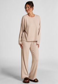 Beige lounge set met een ontspannen lange mouwen top en wijde broek, gemaakt van zachte stof. Het model combineert het met pluizige slippers.