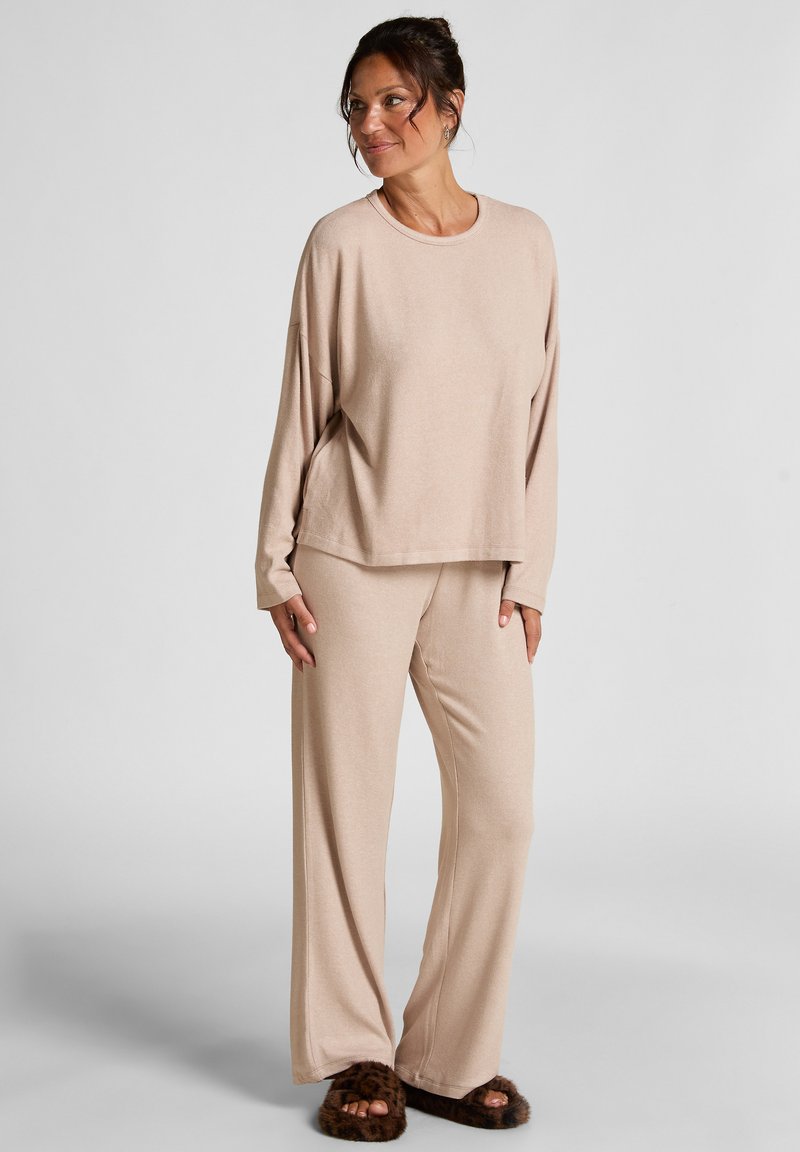 Beige lounge set met een ontspannen lange mouwen top en wijde broek, gemaakt van zachte stof. Het model combineert het met pluizige slippers.