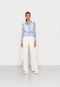 Camisa de botão de manga longa azul claro com padrão floral, combinada com calças largas em creme e sapatos brancos robustos. Pequena mala branca como acessório.