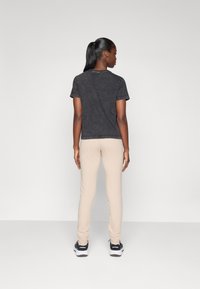 T-shirt en coton noir à manches courtes, associé à un pantalon beige fuselé. Le modèle se tient de dos face à l'appareil photo, portant des baskets noires.