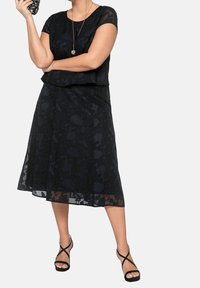Mujer con un vestido de encaje negro con flores, usando tacones negros con tiras y sosteniendo una pequeña cámara negra en su mano levantada.