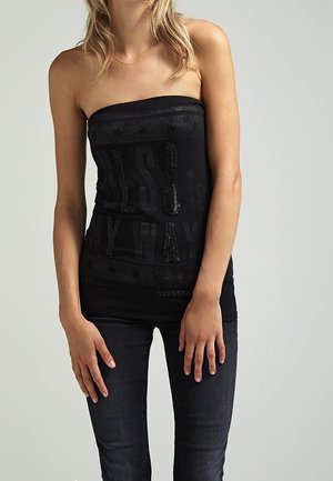 Vrouw draagt een zwarte strapless top met subtiele tekst en donkere spijkerbroek, staand tegen een effen lichtgrijze achtergrond.