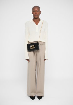 Persona che indossa una maglietta bianca a maniche lunghe, pantaloni beige a gamba larga, scarpe nere a punta e una borsa a tracolla nera "Love Moschino".