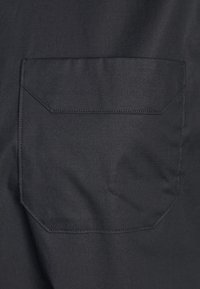 Tela negra con un bolsillo cosido que presenta una forma rectangular y una superficie plana y texturizada. Diseño minimalista sin hardware visible.