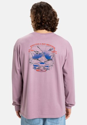 Long-sleeve, pastel purple t-shirt featuring a colorful graphic design on the back with a volcano, palm trees, and the text "Tastes Like Heaven."