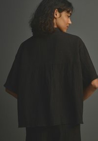 Crna oversized bluza s kratkim rukavima, izrađena od teksturirane tkanine, ravnog kroja i s naboranim dijelom na leđima.