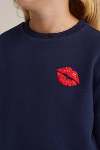 Mornarska dukserica z rdečim vezenim motivom ustnic na levem prsnem prostoru. Tkanina je gladka, ima klasičen kroj s krojenim ovratnikom in rebrastimi manšetami.