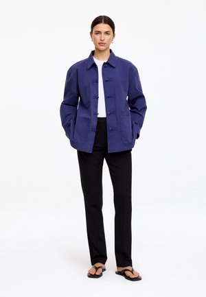 Femme debout portant une veste bleu marine, un haut blanc, un pantalon noir et des sandales noires type tongs sur un fond blanc uni.