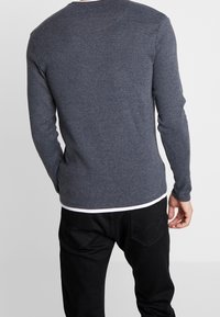 Man wearing a dark gray long-sleeve sweater over a white shirt and black jeans, shown from the back against a plain background.