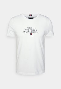 Bílý krátký rukáv tričko s kulatým výstřihem s textem "Tommy Hilfiger MCMLXXXV" a malým červeným, bílým a modrým logem Tommy Hilfiger umístěným na přední straně.