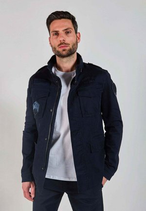 Veste bleu marine avec plusieurs poches, un col haut et un tissu texturé. Dotée d'un logo surdimensionné et d'une fermeture éclair.