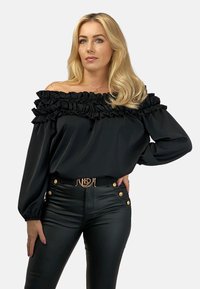 Czarna bluzka typu off-the-shoulder z falbaniastym dekoltem, długimi rękawami i gładką fakturą, zestawiona z czarnymi spodniami z wysokim stanem z materiału sztucznej skóry z złotymi guzikami.