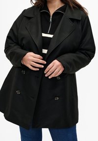 Svart dubbelknäppt rock med stor krage, har en framdragkedja, slät textur och bruna knappar. Bärs över en randig tröja.