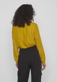 Blouse jaune à motif chevron, avec manches longues et taille élastique, associée à un pantalon cargo noir avec poches latérales.