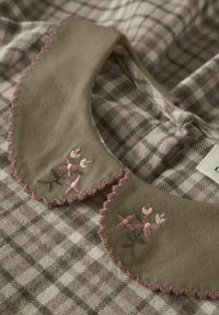 Tissu à carreaux marron et crème avec un col kaki orné de broderies florales roses et de bords festonnés.