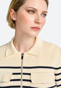 Pletená sveter na zips v krémovej farbe s námorníckymi horizontálnymi pruhmi, s golierom a dvoma náprsníkovými vreckami. Štruktúrovaná tkanina.