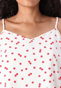 Camisole blanche avec un motif de cerises, fines bretelles et décolleté en V. Le tissu est léger avec une texture douce et des cerises rouges.