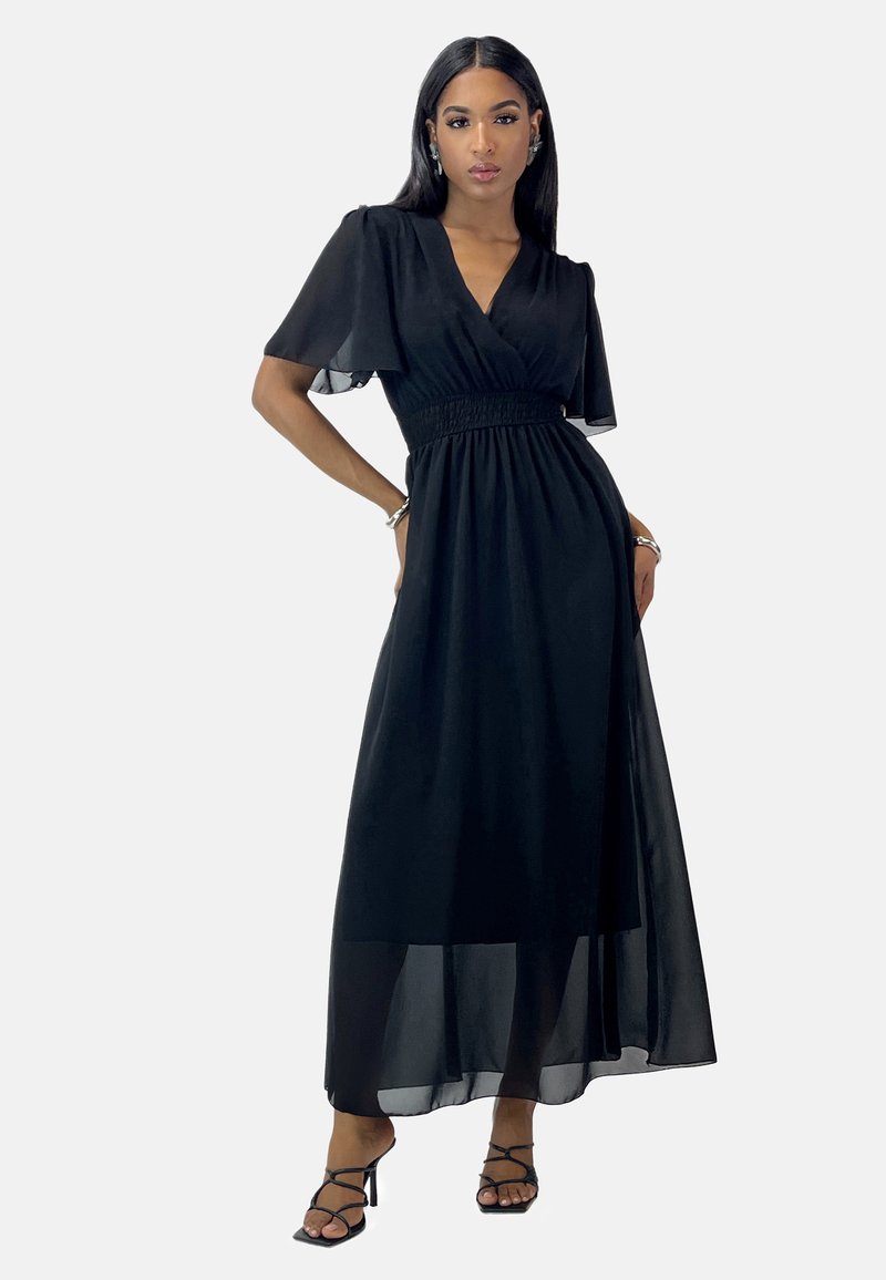 Robe maxi en chiffon noir avec un design enroulé, manches courtes en volants, détail de taille smockée et superposition transparente. Associée à des talons noirs à lanières.
