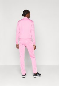 Ροζ φόρμα από απαλό ύφασμα, που περιλαμβάνει ζακέτα με φερμουάρ και ταιριαστό παντελόνι με λευκές ρίγες στα πλάγια. Φοράει μαύρα αθλητικά παπούτσια.