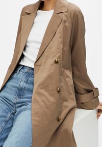 Persona vestida con un abrigo de trench marrón claro sobre una camisa blanca y unos pantalones vaqueros de cintura alta azules, apoyándose en una superficie blanca.