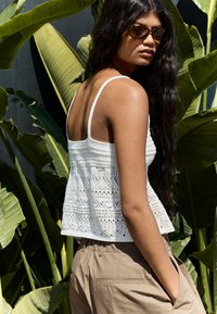 Vit virkad crop top med fina detaljer och justerbara band, tillsammans med beige högmidjade byxor, mot en bakgrund av grön vegetation.
